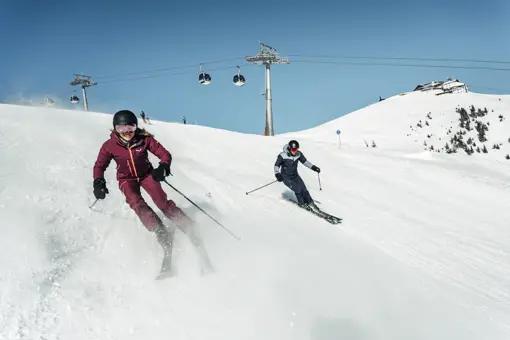 Två skidåkare åker skidor nerför en snötäckt backe i Zell am See, omgivna av alplandskap, en skidort med högsta skidåkning på 3.029 meters höjd.