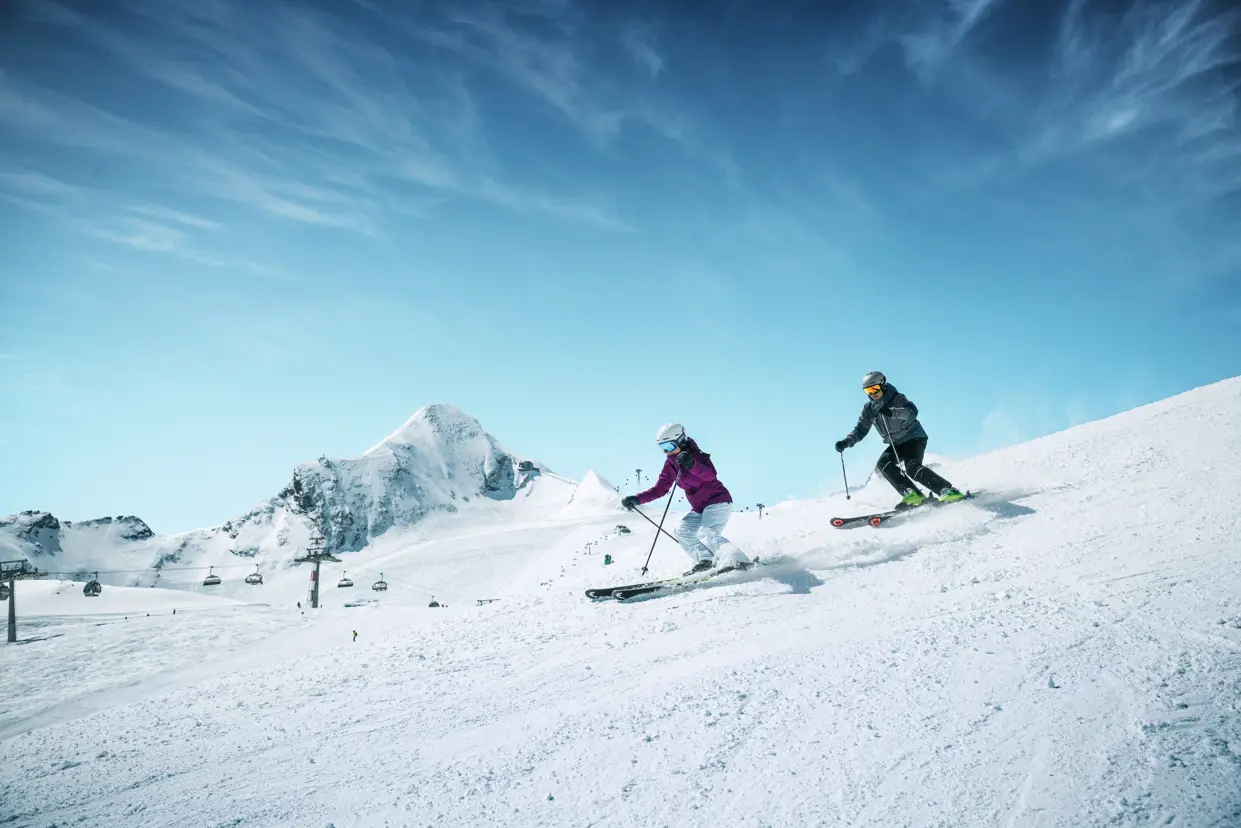 2 vuxna skidåkare åker nerför en snötäckt backe i Zell am See, omgivna av alperna. Perfekta skidförhållanden för vinterälskare, med klar vinterhimmel och vackert alplandskap.