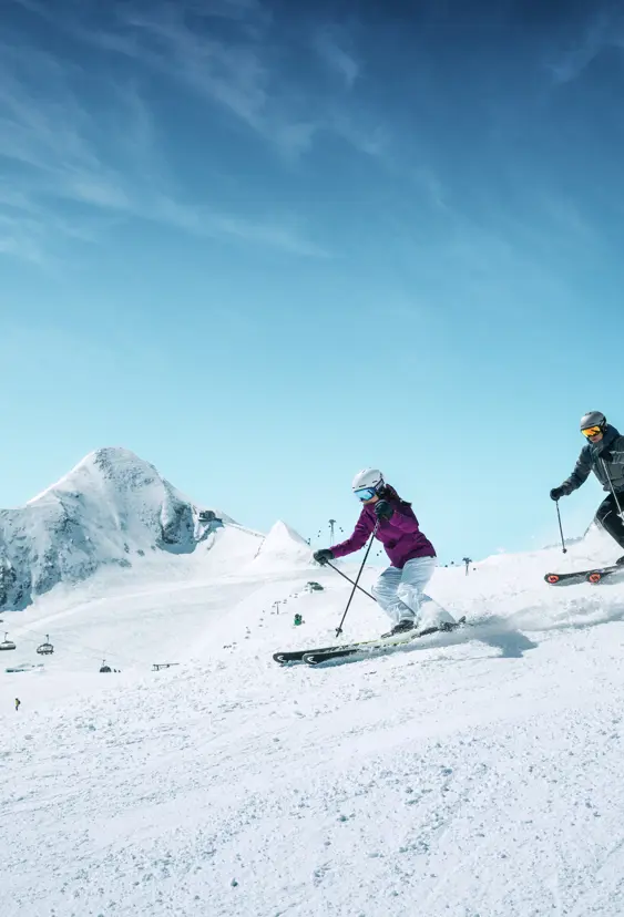 2 vuxna skidåkare åker nerför en snötäckt backe i Zell am See, omgivna av alperna. Perfekta skidförhållanden för vinterälskare, med klar vinterhimmel och vackert alplandskap.