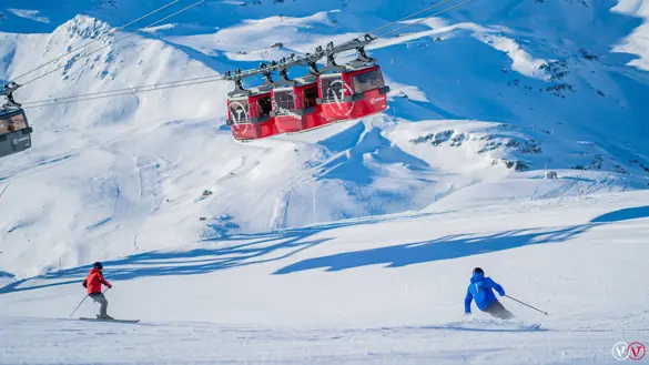 Två skidåkare åker nedför en backe i Val Thorens, nedanför liften, med imponerande utsikt över de snötäckta Alperna. Den omgivande bergskedjan skapar en dramatisk bakgrund medan skidåkarna njuter av perfekt skidåkning i världsklass i ett av Alpernas mest snösäkra områden.