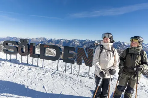Två skidåkare vid Sölden-skylten med snötäckta alptoppar i bakgrunden.