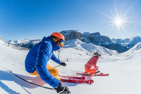 Två skidåkare som åker nerför en pist i Canazei, Dolomiti Superski som är ett av världens största skidområde, omgiven av sol, snötäckta berg och fantastisk utsikt.
